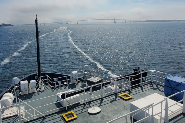 The back deck of a ship sailing on blue water with the wake in the background on a cloudy day. A suspension bridge is visible in the distance.