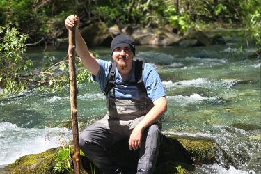 A person wearing waders sits next to a river holding a large branch