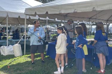 A white man in a blue button down shirt and shorts standing next to a tent holds a fishing reel while several children watch.
