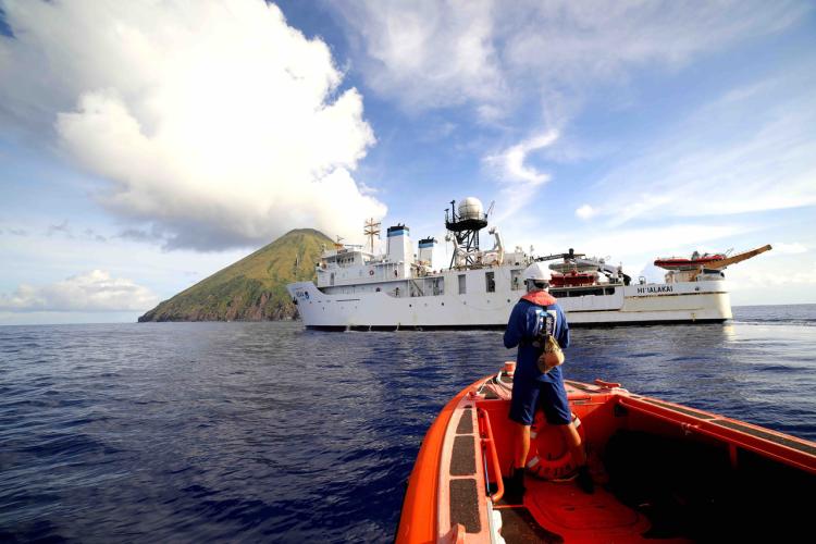A small orange boat in the foreground with a person standing at the front looking at a white NOAA ship and small island ahead in the background.