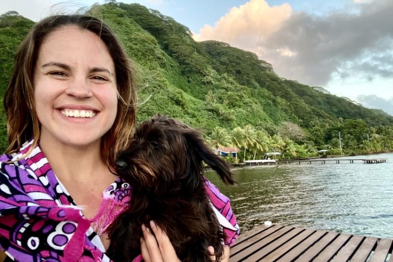 A woman smiling and holding a small black dog in front of green mountains and the ocean.