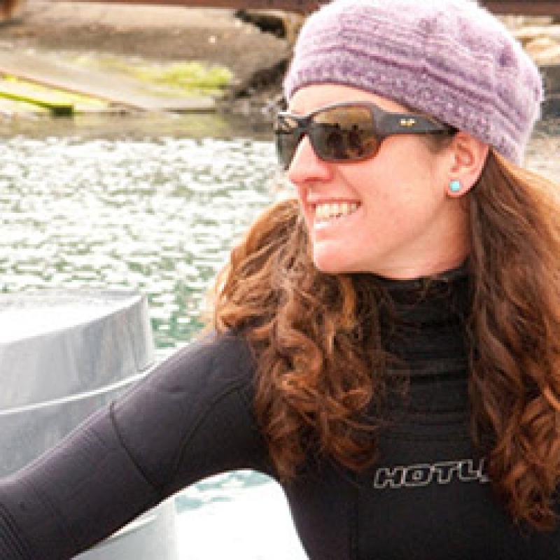 Portrait of scientist wearing a hat, glasses and black turtleneck with water in the background.