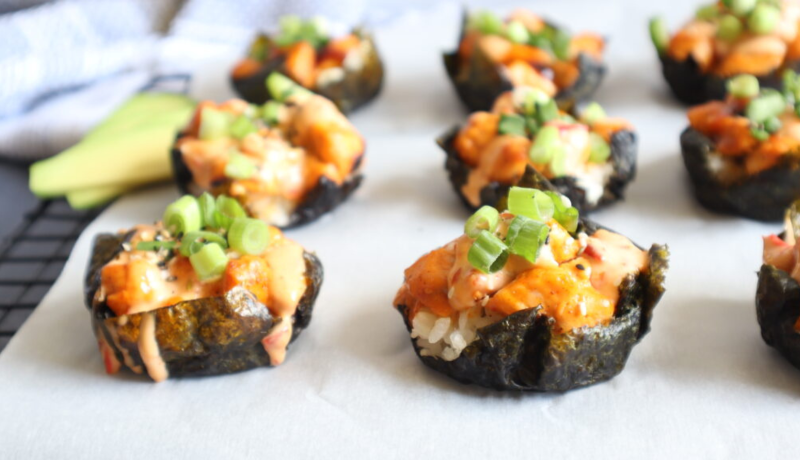rice, salmon, an orange dressing, and sliced chives arranged in a cup made out of dried seaweed sheets. the cups are arranged in three rows paced on white parchment paper.